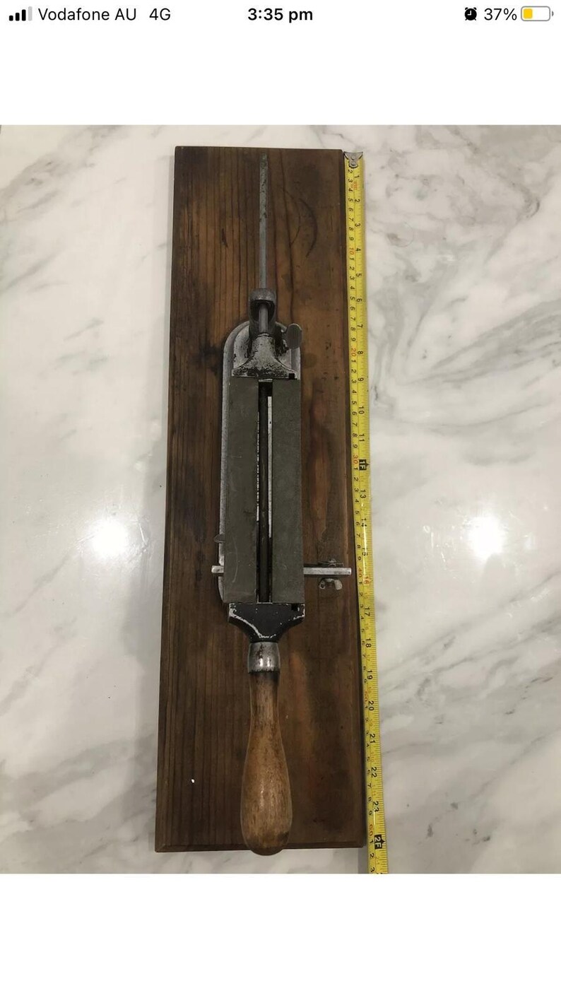May include: An antique knife sharpener mounted on a wooden base. The sharpener is made of metal with a wooden handle. A yellow measuring tape is placed next to the sharpener, showing a length of approximately 24 inches.