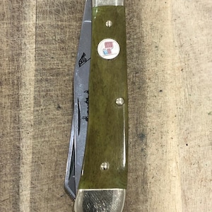 Op de afbeelding: Een zakmes met een groene handgreep en een zilveren lemmet. De handgreep heeft een cirkelvormig embleem met een rood-wit ontwerp. Het lemmet heeft een zwarte afbeelding van een zebra. Het mes ligt op een houten oppervlak.