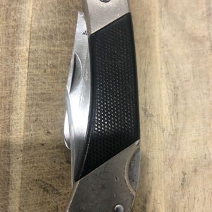 Puede incluir: Una navaja de bolsillo plegable con un mango negro texturizado y una hoja plateada. La navaja tiene un refuerzo plateado y una parte superior plateada. La hoja est&aacute; parcialmente abierta, revelando el filo. La navaja est&aacute; cerrada y lista para usar.
