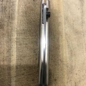 May include: A closed, silver folding knife with a black screw. The knife is made of metal and has a smooth, reflective surface. The blade is not visible in this image. The knife is resting on a wooden surface.