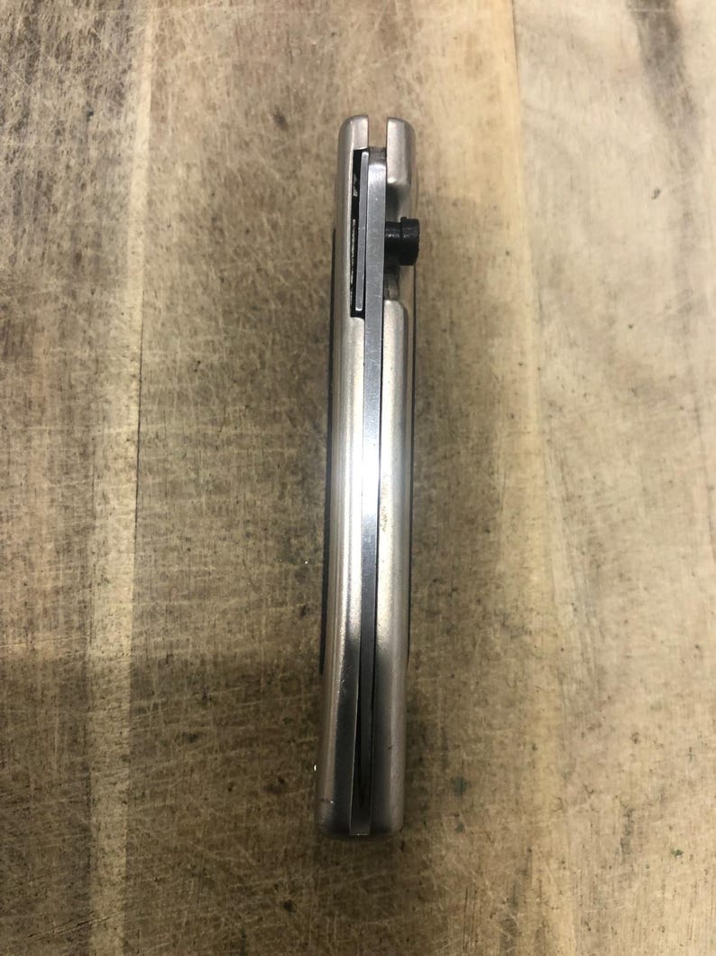May include: A closed, silver folding knife with a black screw. The knife is made of metal and has a smooth, reflective surface. The blade is not visible in this image. The knife is resting on a wooden surface.