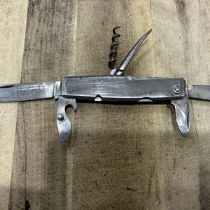 May include: A silver-toned multi-tool pocket knife featuring a main blade, a smaller secondary blade, a corkscrew, a bottle opener, and a flathead screwdriver. The metal handle shows signs of wear with visible scratches and a matte finish.
