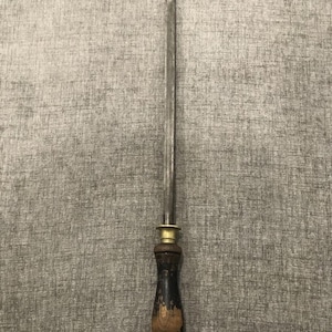May include: A steel knife sharpening steel with a wooden handle. The handle is dark brown and black with a brass collar. The steel is long and slender, designed for sharpening knives. A small metal loop is attached to the bottom of the handle.