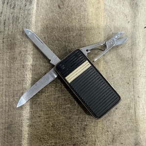 May include: A multi-tool pocket knife featuring a rectangular black and white ribbed handle. The tool includes a stainless steel blade, a metal nail file, and a pair of small scissors extended outward. The blade is stamped with the text Stainless Steel Japan.