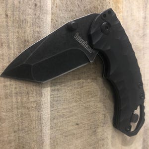 May include: A black Kershaw folding knife with a textured handle and a partially open blade. The blade is angled and has a sharp edge. The word "Kershaw" is printed on the blade. The knife is set against a light brown wooden surface.