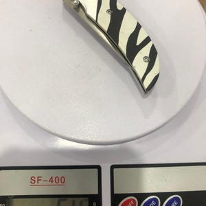 K&ouml;nnte beinhalten: Ein Klappmesser mit Zebrastreifenmuster auf dem Griff und einem rosa Logo. Das Messer liegt auf einer Waage, die 64 oz anzeigt.