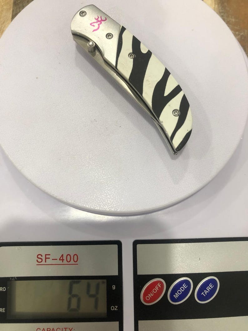 K&ouml;nnte beinhalten: Ein Klappmesser mit Zebrastreifenmuster auf dem Griff und einem rosa Logo. Das Messer liegt auf einer Waage, die 64 oz anzeigt.