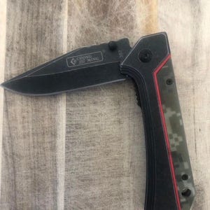 May include: A black folding knife with a partially open blade. The knife features a black blade with the text "CHONG MING" and a handle with a digital camouflage pattern and red accents. The knife is on a wooden surface.