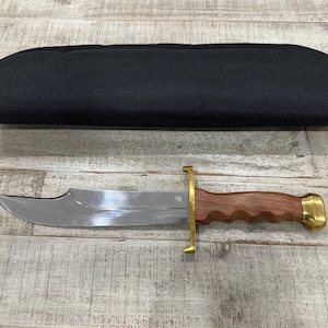 May include: A fixed-blade knife with a polished silver steel blade, a brass crossguard, and a contoured brown wooden handle with a brass pommel. The knife is accompanied by a black fabric sheath with a zipper closure along the top edge.