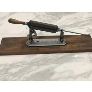 May include: Antique knife sharpener with a wooden handle and a metal blade. The sharpener is mounted on a wooden base and has a metal support structure. The overall dimensions are approximately 12 inches long and 4 inches wide.