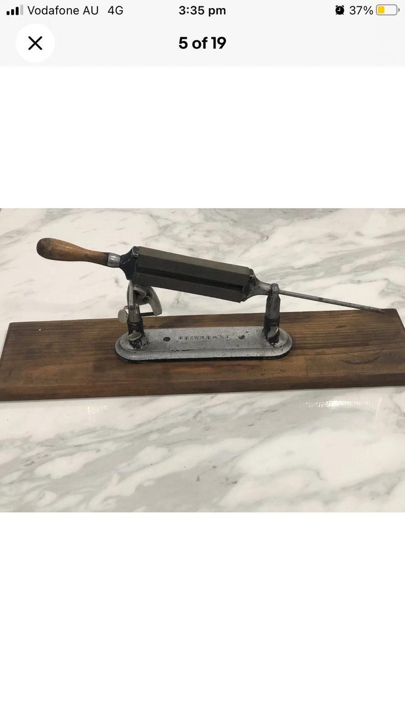 May include: Antique knife sharpener with a wooden handle and a metal blade. The sharpener is mounted on a wooden base and has a metal support structure. The overall dimensions are approximately 12 inches long and 4 inches wide.