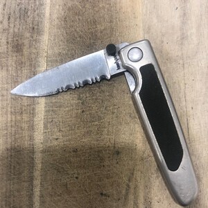 May include: A partially open folding knife with a serrated blade. The blade is silver, and the handle is silver with a black rubber grip. The knife has the text "Kershaw" on the blade.