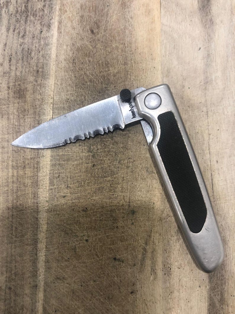 May include: A partially open folding knife with a serrated blade. The blade is silver, and the handle is silver with a black rubber grip. The knife has the text "Kershaw" on the blade.