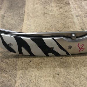 K&ouml;nnte beinhalten: Ein Klappmesser mit einer Klinge und einem Griff aus Edelstahl. Der Griff hat ein schwarz-wei&szlig;es Zebrastreifenmuster. Das Messer hat einen silberfarbenen Metallrahmen und ein rosa Logo.
