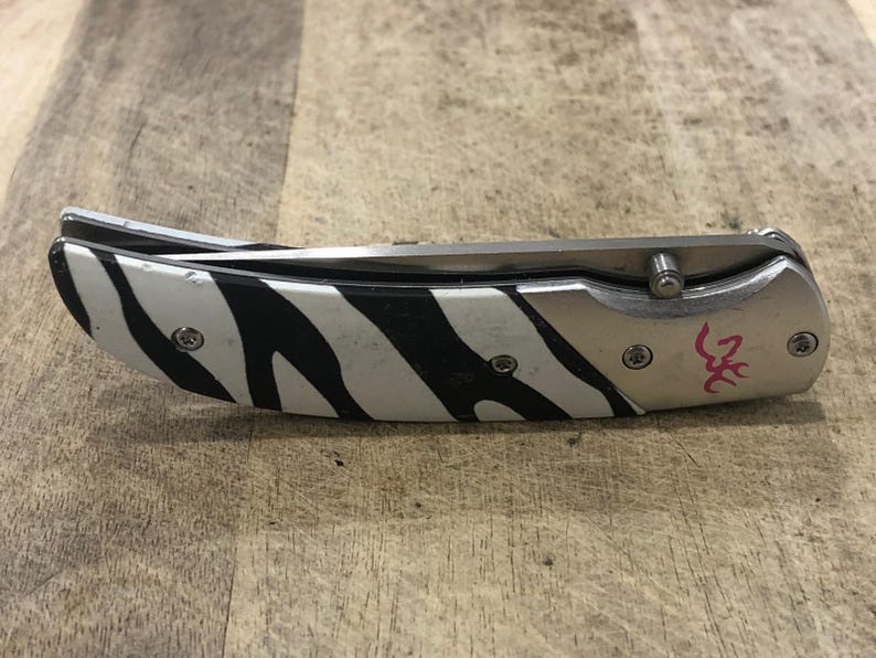 K&ouml;nnte beinhalten: Ein Klappmesser mit einer Klinge und einem Griff aus Edelstahl. Der Griff hat ein schwarz-wei&szlig;es Zebrastreifenmuster. Das Messer hat einen silberfarbenen Metallrahmen und ein rosa Logo.