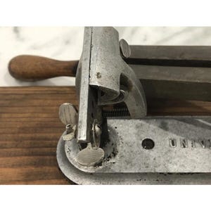 May include: Close-up of a vintage metal tool with a wooden handle. The tool has a silver finish and features adjustable parts, including a screw and a circular base. The word "UNIV" is visible on the base.