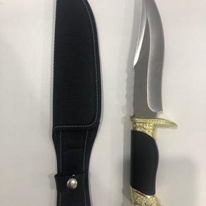 Pode incluir: Uma faca de caça em aço inoxidável com cabo preto e detalhes dourados, ao lado de uma bainha preta. A lâmina da faca é afiada e curva. A pega apresenta um design dourado decorativo.