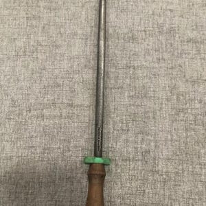 May include: A steel knife sharpening rod with a wooden handle and a green plastic guard. The rod is long and slender, designed for honing the edge of knives. The handle is a dark brown color.