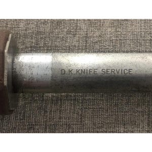 Pode incluir: Close-up de uma l&acirc;mina de faca de metal com o texto "D.K. KNIFE SERVICE" gravado na superf&iacute;cie. A l&acirc;mina tem uma apar&ecirc;ncia desgastada e industrial, sugerindo uso frequente. O fundo &eacute; uma superf&iacute;cie texturizada de tons neutros.
