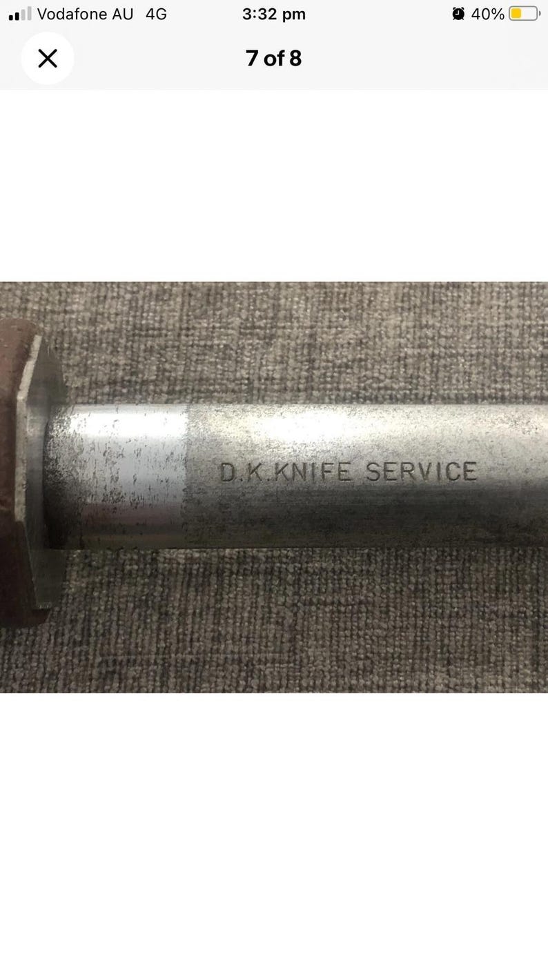 Pode incluir: Close-up de uma l&acirc;mina de faca de metal com o texto "D.K. KNIFE SERVICE" gravado na superf&iacute;cie. A l&acirc;mina tem uma apar&ecirc;ncia desgastada e industrial, sugerindo uso frequente. O fundo &eacute; uma superf&iacute;cie texturizada de tons neutros.