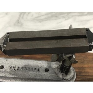 May include: A vintage metal tool with the word "UNIVERSAL" stamped on the base. The tool has a dark, rectangular top section with two parallel grooves. The base is silver-colored, and the tool appears to be mounted on a wooden surface.