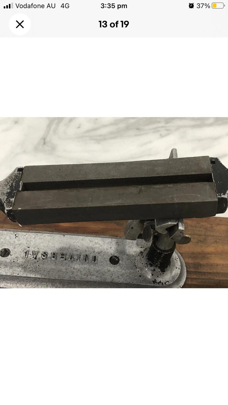 May include: A vintage metal tool with the word "UNIVERSAL" stamped on the base. The tool has a dark, rectangular top section with two parallel grooves. The base is silver-colored, and the tool appears to be mounted on a wooden surface.