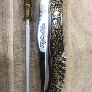 May include: A silver knife sharpener with a brass handle next to a folding knife. The knife has a brass-colored handle with a fish and floral design. The blade has the text "Fighter Plus".