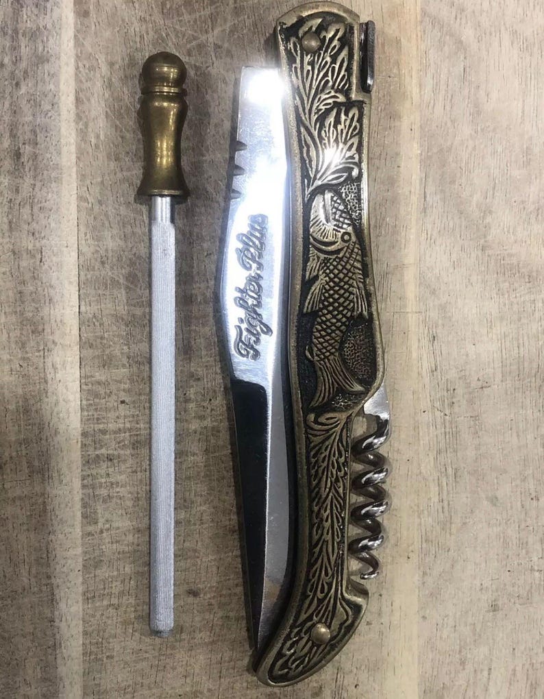 May include: A silver knife sharpener with a brass handle next to a folding knife. The knife has a brass-colored handle with a fish and floral design. The blade has the text "Fighter Plus".