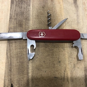 Puede incluir: Una navaja suiza roja con múltiples herramientas desplegadas, incluyendo una hoja, un abridor de botellas y un sacacorchos. La navaja presenta el icónico emblema de la cruz blanca. Las herramientas son de metal plateado.