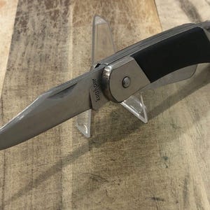 Puede incluir: Una navaja plegable plateada y negra con una hoja parcialmente dentada. El cuchillo tiene un mango de goma negro y un marco de metal plateado. La hoja est&aacute; en &aacute;ngulo y tiene el texto "Kershaw". El cuchillo se exhibe en un soporte de pl&aacute;stico transparente.