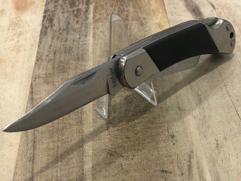 Puede incluir: Una navaja plegable plateada y negra con una hoja parcialmente dentada. El cuchillo tiene un mango de goma negro y un marco de metal plateado. La hoja est&aacute; en &aacute;ngulo y tiene el texto "Kershaw". El cuchillo se exhibe en un soporte de pl&aacute;stico transparente.