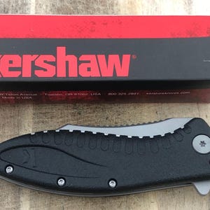 Peut inclure: Un couteau pliant noir et gris avec un manche texturé et une lame argentée. Le couteau est à côté d'une boîte rouge et noire avec le nom de la marque "Kershaw" en lettres rouges. La boîte contient également l'adresse et le site Web.