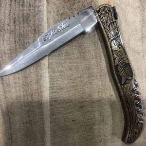 May include: A stainless steel folding knife with the text "Fighter Plus" etched on the blade. The handle is gold-toned with an engraved fish and floral design. The knife is open, displaying the sharp blade.