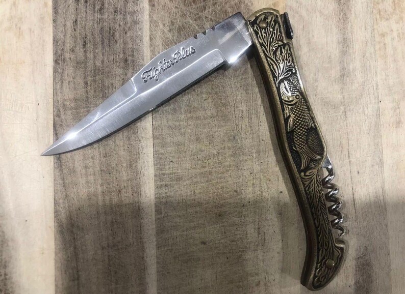 May include: A stainless steel folding knife with the text "Fighter Plus" etched on the blade. The handle is gold-toned with an engraved fish and floral design. The knife is open, displaying the sharp blade.