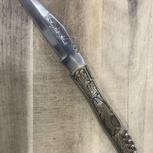 May include: A stainless steel knife with a decorative handle featuring a fish and leaf design. The blade has the text "Fisher Club" etched on it. The knife is set against a wooden background.