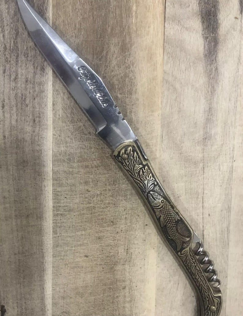 May include: A stainless steel knife with a decorative handle featuring a fish and leaf design. The blade has the text "Fisher Club" etched on it. The knife is set against a wooden background.