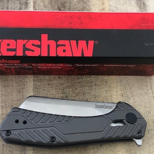 Peut inclure: Un couteau pliant Kershaw gris avec une lame argentée, posé sur une surface en bois. Le couteau a un manche texturé et le logo Kershaw est visible. Une boîte Kershaw rouge et noire est en arrière-plan.