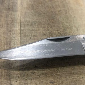 Puede incluir: Primer plano de una hoja de navaja plegable plateada. La hoja es afilada y tiene la palabra "Kershaw" grabada. La navaja est&aacute; parcialmente abierta, revelando la punta. El fondo es una superficie de madera.