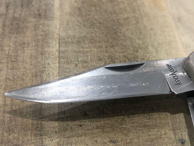 Puede incluir: Primer plano de una hoja de navaja plegable plateada. La hoja es afilada y tiene la palabra "Kershaw" grabada. La navaja est&aacute; parcialmente abierta, revelando la punta. El fondo es una superficie de madera.