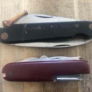 Puede incluir: Dos navajas de bolsillo sobre una superficie de madera. La navaja superior tiene un mango negro con el texto "SHERMAN BRAND" y una hoja plateada. La navaja inferior tiene un mango granate y una hoja plateada.