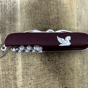 May include: A maroon multi-tool pocket knife featuring a silver Pegasus logo on the handle. The tool includes a metal key ring attached to one end and visible metal components such as a corkscrew and blade edges along the side of the casing.