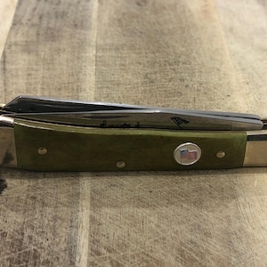 Op de afbeelding: Een gesloten zakmes met een groene handgreep en zilveren messen. De handgreep heeft een klein Amerikaans vlagembleem en messingkleurige metalen accenten. De messen zijn gedeeltelijk zichtbaar. Het mes ligt op een houten oppervlak.