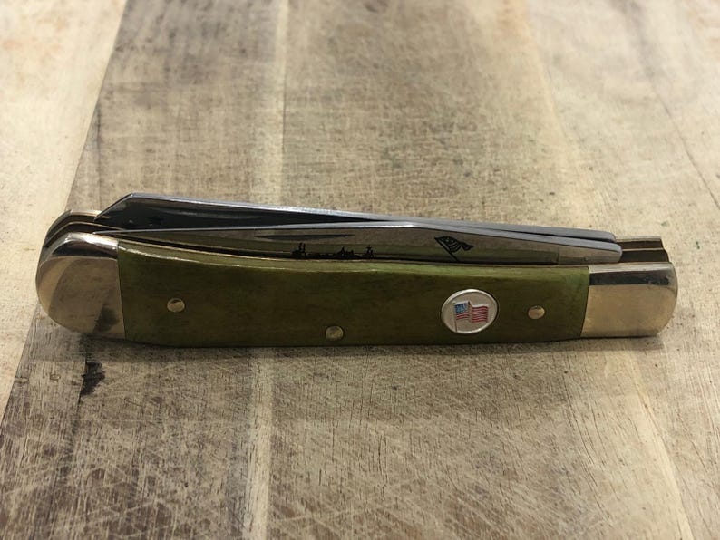 Op de afbeelding: Een gesloten zakmes met een groene handgreep en zilveren messen. De handgreep heeft een klein Amerikaans vlagembleem en messingkleurige metalen accenten. De messen zijn gedeeltelijk zichtbaar. Het mes ligt op een houten oppervlak.