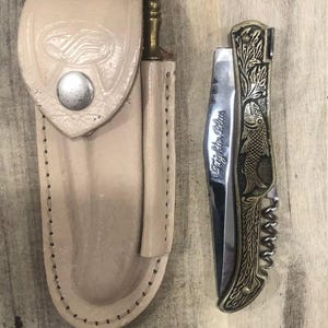 May include: A beige leather knife sheath with a silver snap closure next to a decorative folding knife. The knife has a silver blade with the words "Fightin' Plus" and a gold-colored handle with an engraved fish design.