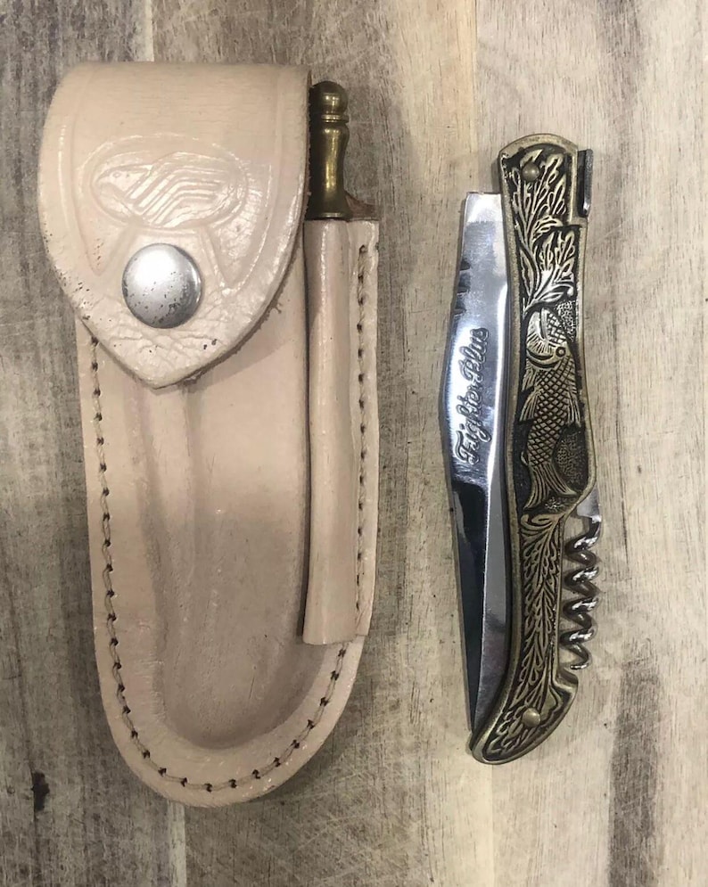 May include: A beige leather knife sheath with a silver snap closure next to a decorative folding knife. The knife has a silver blade with the words "Fightin' Plus" and a gold-colored handle with an engraved fish design.