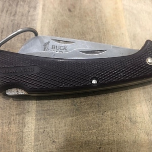 May include: A black and silver folding knife with a textured handle and a metal clip. The blade is partially open, displaying the word "BUCK" and a logo. The handle has the words "BUCK WORK-MAN" printed on it.