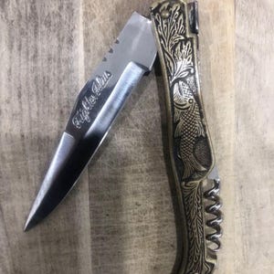 May include: A partially open pocket knife with a silver blade and a decorative brass handle. The blade is engraved with the text "Fighter Plus". The handle features an etched fish design and a corkscrew.