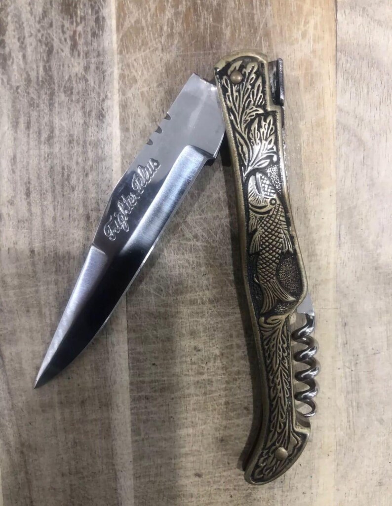 May include: A partially open pocket knife with a silver blade and a decorative brass handle. The blade is engraved with the text "Fighter Plus". The handle features an etched fish design and a corkscrew.