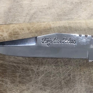May include: Close-up of a stainless steel knife with the text "Fighter Plus" etched on the blade. The knife has a sharp point and a decorative handle, set against a wooden surface.
