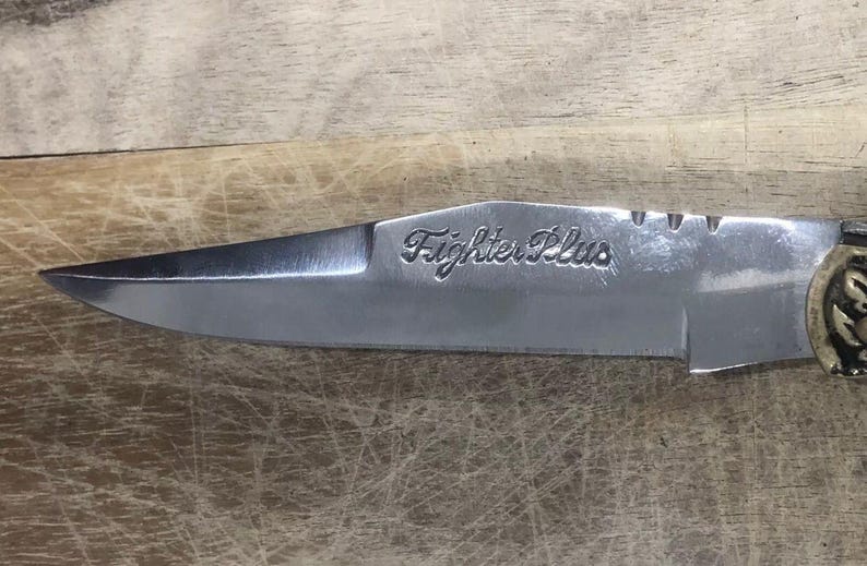May include: Close-up of a stainless steel knife with the text "Fighter Plus" etched on the blade. The knife has a sharp point and a decorative handle, set against a wooden surface.